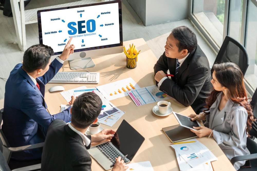 Group of professionals conducting a B2B SEO audit in a boardroom with a computer screen displaying SEO-related terms such as keywords, content, social media, and link."